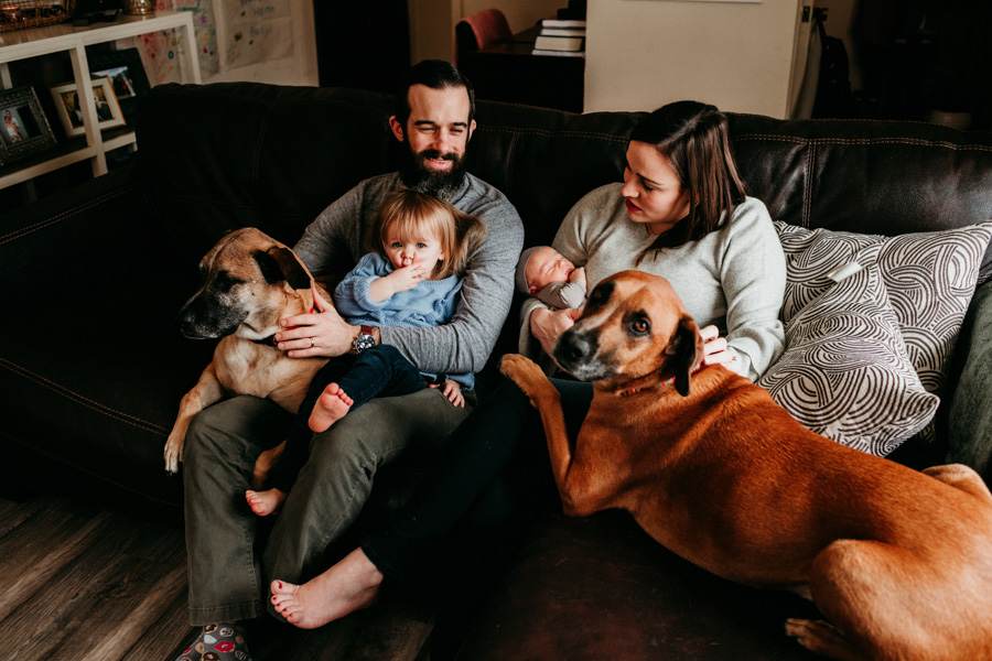 A family snuggling on the couch with their dogs and two kids, baby sleeping while toddler makes a silly face